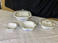 Front angled view of 4 nested white Corningware baking dishes with blue floral pattern and glass lids on largest and medium sized dishes, showing size gradation and overall condition.