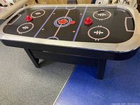 Full view of the black air hockey table showing the playing surface, red mallets, black and orange pucks, and silver trim.