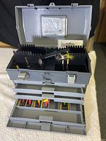 Open grey plastic fishing tackle box showing three drawered trays containing assorted colorful fishing lures. Waterproof compartment inside lid with slots for license storage.