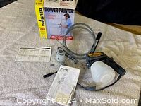 Electric paint sprayer with attached tubing, paint container, instruction manuals, and original box on beige fabric surface.