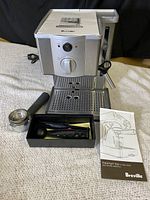 Front view of Breville Café Roma espresso machine with portafilter, black box of accessories, and instruction manual on countertop.