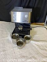 Front view of Via Venezia espresso machine with three stainless steel measuring cups on a cloth surface, control buttons visible on the machine front.