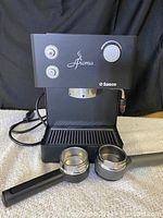 Full front view of the black Saeco Aroma espresso machine, showing buttons, logo, and one portafilter attached plus one placed alongside the machine.