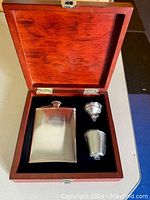 Open wooden case showing polished metal flask, shot glass, and jigger inside black lining