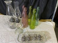 Two clear hurricane lamps, six colored textured glass bottles in varying colors, and a rectangular clear glass serve tray with six metal violin-shaped napkin rings arranged on a beige carpet.