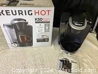 Keurig K50 Hot coffee maker with the original box and instructions displayed. Coffee maker is unpackaged and positioned alongside the box on a light carpet.