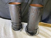 Two cylindrical metal lamp diffusers with black tree branch cutout pattern and bronze-colored inside, each with power cord