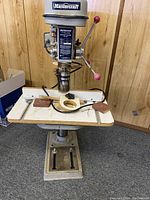 Front view of the Mastercraft drill press showing the control lever with red knobs, drill chuck, and work surface with wooden inserts.