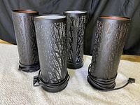 Four black cylindrical lamps or oil diffusers displayed side by side, showing their exterior tree branch cut-out pattern and attached cords.