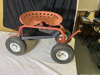 Side view of red metal mobile gardener stool with large knobby tires and black tool tray under the adjustable tractor-style seat.