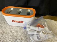 ZOKU fast popsicle maker with orange trim, white and orange plastic, with multiple white popsicle sticks and orange molds in a sealed plastic bag beside it.