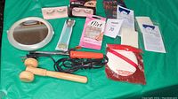 Photo showing assorted beauty aids including curling iron, wooden massager hammer, false eyelashes, callus shaver, waxing strips, mirror and nail accessories on green background.