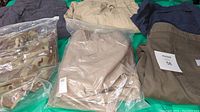 Wide shot of the lot showing multiple pants and shorts some sealed in plastic, including camo and tan pants and various folded pants with size 44 and 42 tags.