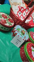 Four round Christmas-themed cookie tins in red, green and blue with holiday motifs, placed on green surface next to red woven basket with gift bags.