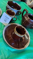 Two brown drip glaze saucers and three matching cups displayed with a brown teapot on a green cloth surface.