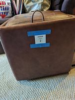 Side and bottom view of the brown leather storage ottoman showing some wear and a fabric handle attached to top.