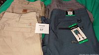 Four men's pants and shorts laid out on green background. Pants are beige, gray, and light beige Carhartt, and blue/gray O'Neill shorts with tags.