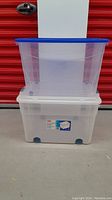 Two clear plastic tote bins stacked, showing size and design with blue lids and wheels.
