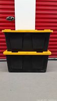 Front view of two black industrial tote bins stacked, showing sturdy yellow lids and ribbed sides