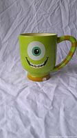Mike Wazowski green ceramic mug with single large eye, smiling mouth, striped handle, and small chip on the base visible.