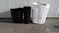 Lot of 3 bins including 1 white laundry basket and 2 black plastic bins placed on pavement outside.