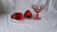 Lot of 3 red blown glass pieces displayed on white cloth: a swan dish on left, a heart-shaped bowl center, and a ruffled vase right.