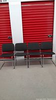 Four stackable metal chairs with dark upholstered seats and backs, arranged side by side, photographed in front of a red storage door.