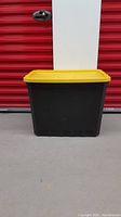 Front view of tall, black Sterilite tote bin with yellow lid closed, showing rectangular shape and height.