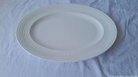 Oval white bone china serving plate with embossed decorative rim detail, placed on a white fabric background.
