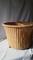 Side and angled view of a handwoven wicker laundry basket with small black feet at the base, showing the basket against a white fabric backdrop.