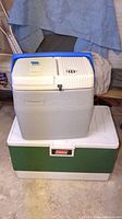 Green Coleman cooler under a grey electric Coleman cooler showing hinged lids and red Coleman logos, with visible stains and wear.