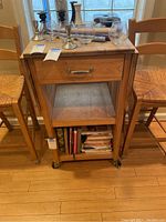 Front view of the wooden chef's cart showing the drawer, open middle shelf with some items, and bottom storage with books, flanked by two wooden chairs.