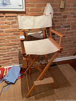 Two wooden director's chairs with beige canvas seat and back, natural wood frame, armrests, and footrest. Chairs placed in front of a brick wall and carpeted floor.