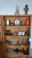 Wooden shelf unit displaying a mix of items including a metallic pitcher, orange ceramic containers, a small birdhouse tissue box cover, pottery bowls, and figurines including a Native American kachina doll.