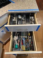 Kitchen drawer with mesh organizers holding assorted stainless steel flatware, black handled cooking utensils, white plastic spoons, wine stoppers, and various kitchen tools.