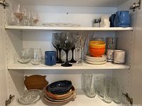 Shelf view with various glassware including wine glasses, tumblers, mugs, stacked bowls, ceramic pitchers, and two wooden cutting boards