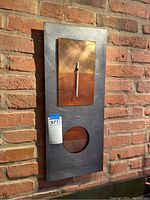Front and angled views of the rectangular metal wall clock showing brushed steel surface, copper-tone clock face with silver hands, and circular window at bottom.