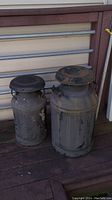 Two black metal milk jugs sitting outside on wooden deck with metal railings behind. Jugs show significant weathering and rust.