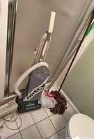 Standing Oreck vacuum with fabric bag, mop handles, and cleaning supplies on tile floor.