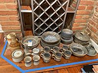 Wide view of assorted pottery dishware including plates, bowls, mugs, jars, and pottery pitchers arranged on a wooden surface against a brick wall.
