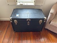 Front diagonal view of a black vintage trunk with metal corner protection, lock, and clasps on top.