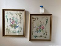 Photo of two framed needlepoint floral artworks hanging on a wall, showing full view of both frames and embroidery designs.
