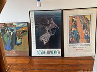 Three framed pieces of wall art leaning against a wall: Picasso-themed print on left, Sonora Mexico aerial photo in center, Brightest London Underground poster on right.