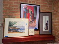 Four framed artworks displayed on a shelf against brick wall, showing different sizes and styles of landscape-themed art