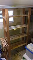Four-tier wooden shelf unit constructed of raw wood slats and thick vertical planks, located inside a basement.