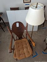 Wooden antique style rocking chair with curved armrests and rounded backrest, tufted removable tan cushions on seat and back, alongside a floor lamp with tall slender brass pole and cream lampshade.