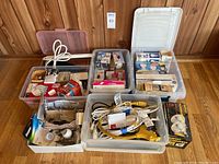 Overview of multiple plastic bins containing lightbulbs, extension cords, electrical outlets, batteries, and other small electrical items arranged on a wooden floor with wood-paneled wall background.