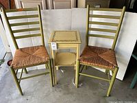 Pair of green ladder-back chairs with woven rattan seats flanking a small yellow occasional table with gold painted vine decorations.