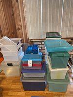 Stacked assortment of plastic storage bins in clear, light blue, green, and other colors, arranged on wood floor near a window.