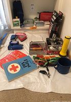 Wide view of assorted emergency and survival gear spread on table including first aid books, flashlights, knives, utility pouches, cords, whistles, and a blue camping mug.
